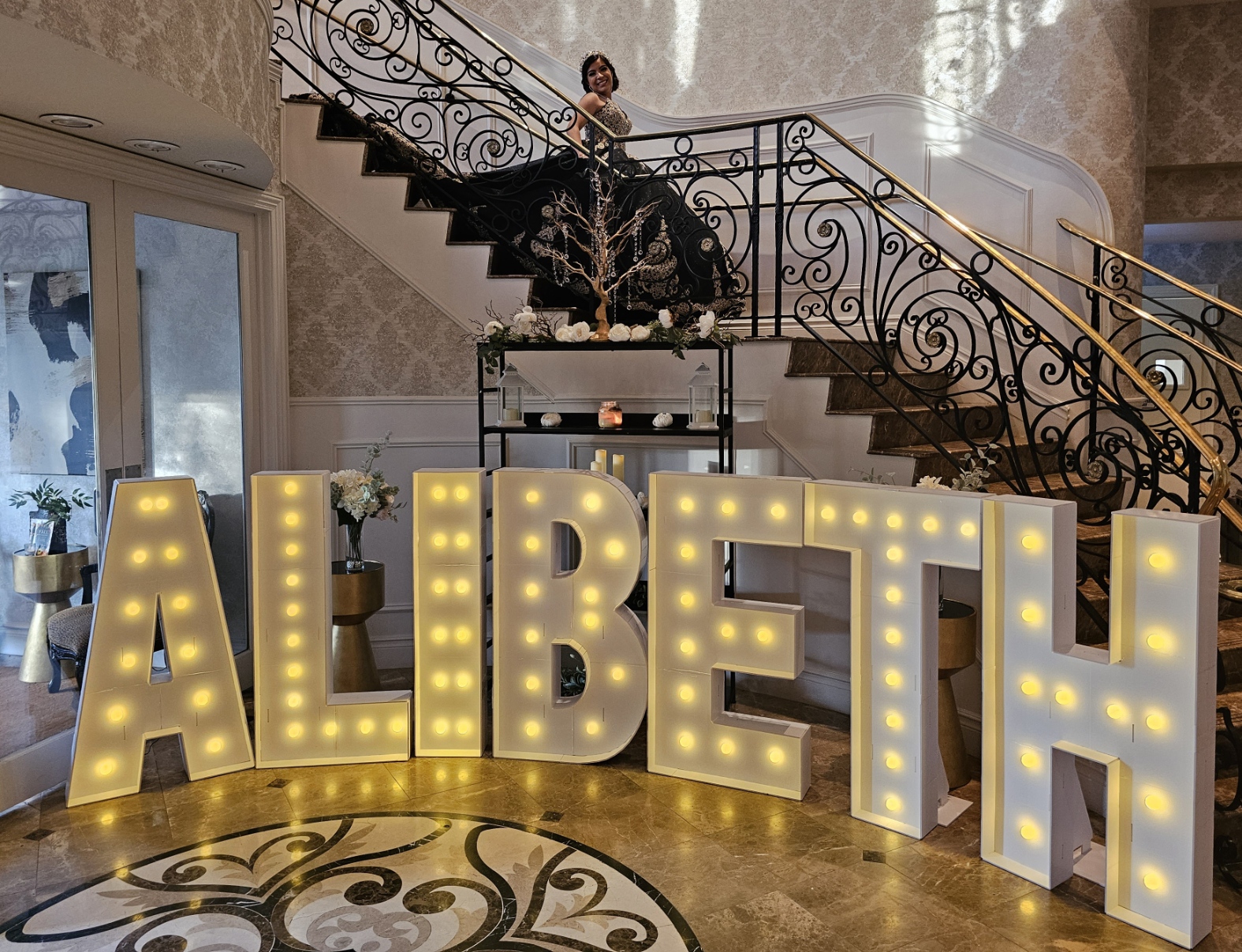 Alibeth's Quinceanera: Alibeth walking down a grand staircase and 4ft tall marquee letters that spell out ALIBETH with warm lights in the front at the Vanderbilt in Staten Island, New York done by PMT Party Services Group